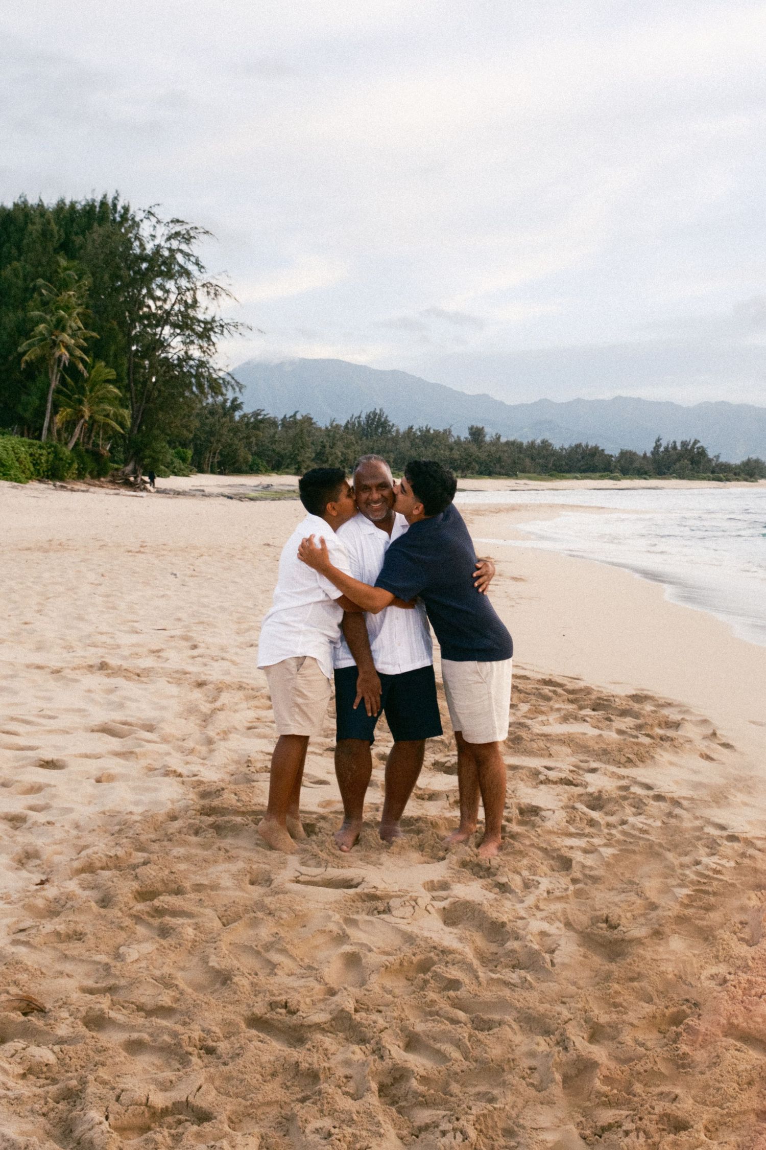 Family of four walking hand-in-hand along a sandy beach at dusk with mountains in the background.