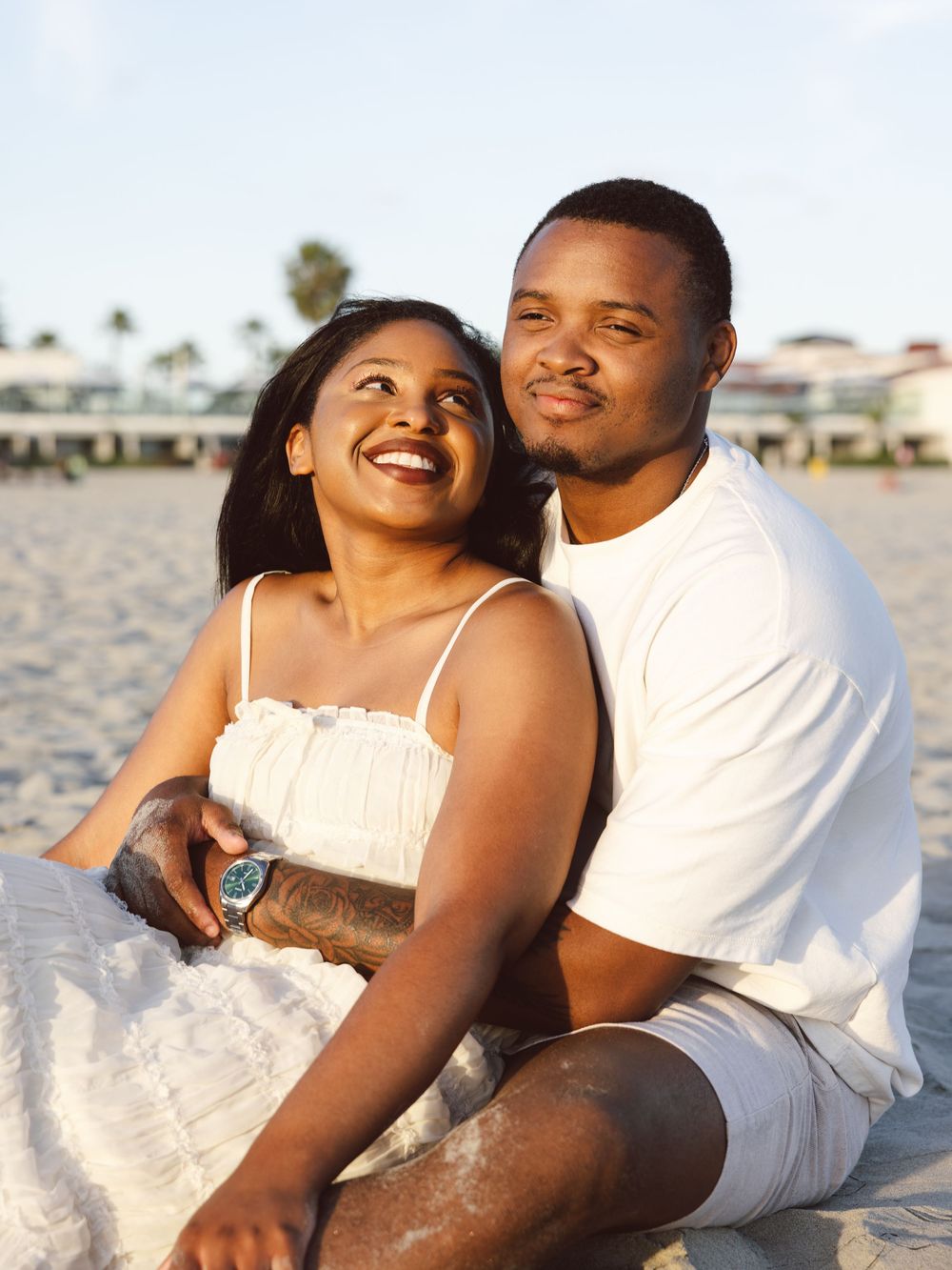 Happy couple sitting together on a sandy beach, wearing white outfits, smiling and enjoying a sunny day outdoors.