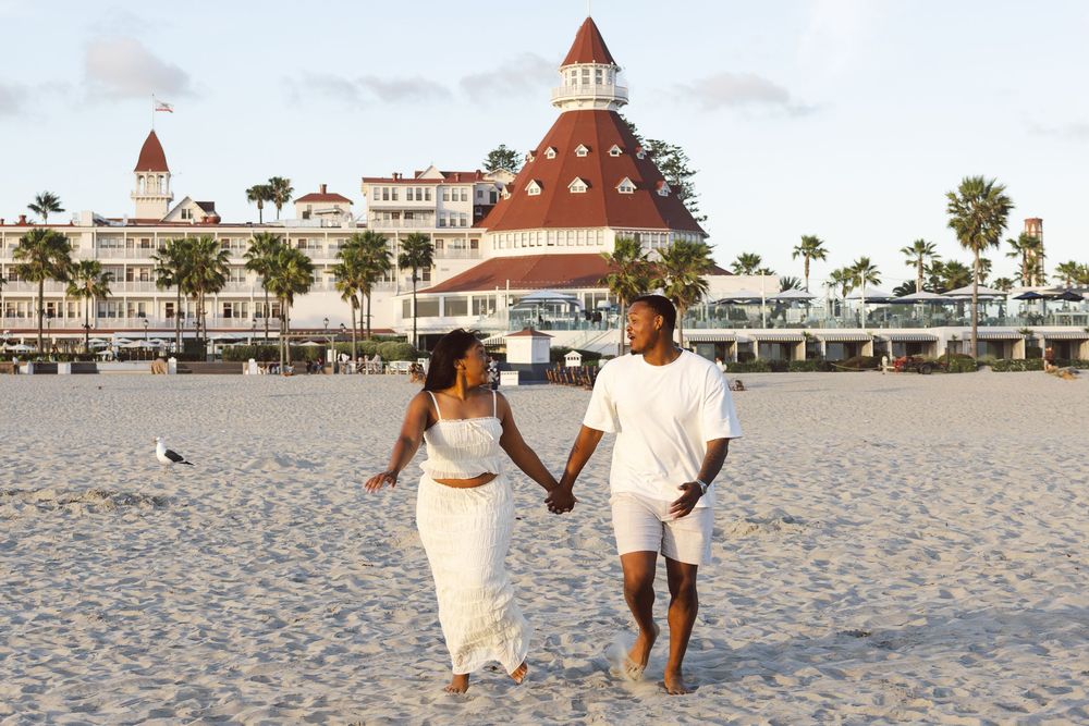 A couple holds hands walking on a sandy beach in front of the historic Hotel del Coronado in San Diego, California.