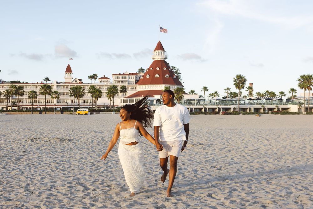 Couple in white outfits running on sandy beach with Hotel del Coronado in background on a clear day.