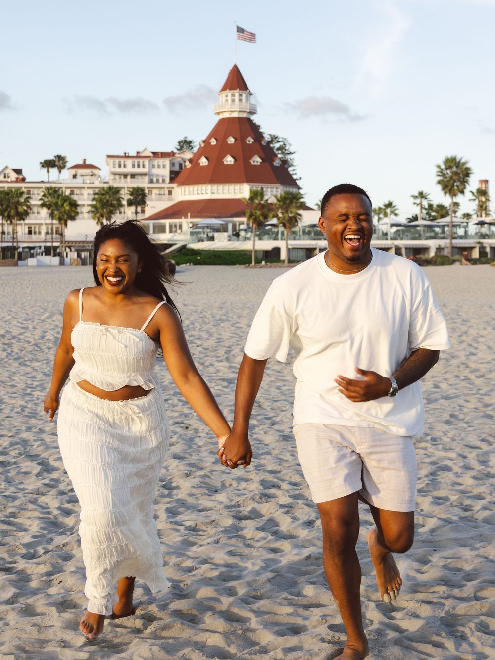 A joyful couple in matching white outfits runs hand-in-hand on a sandy beach near the Hotel del Coronado.