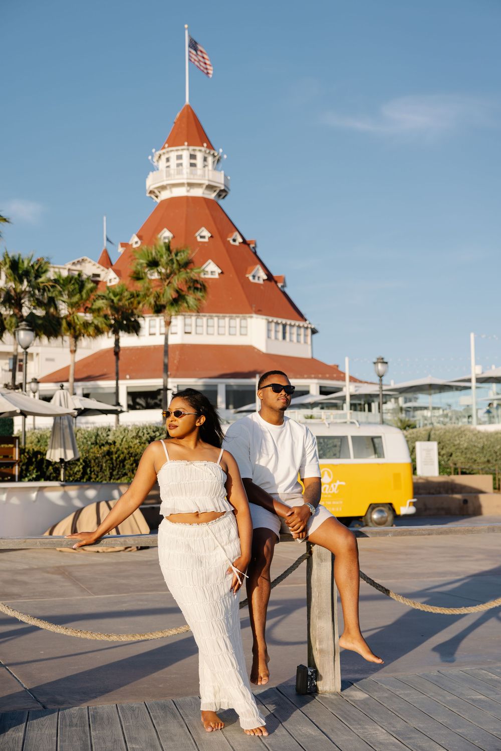 Couple in white outfits posing near Hotel del Coronado with palm trees and American flag on sunny day.