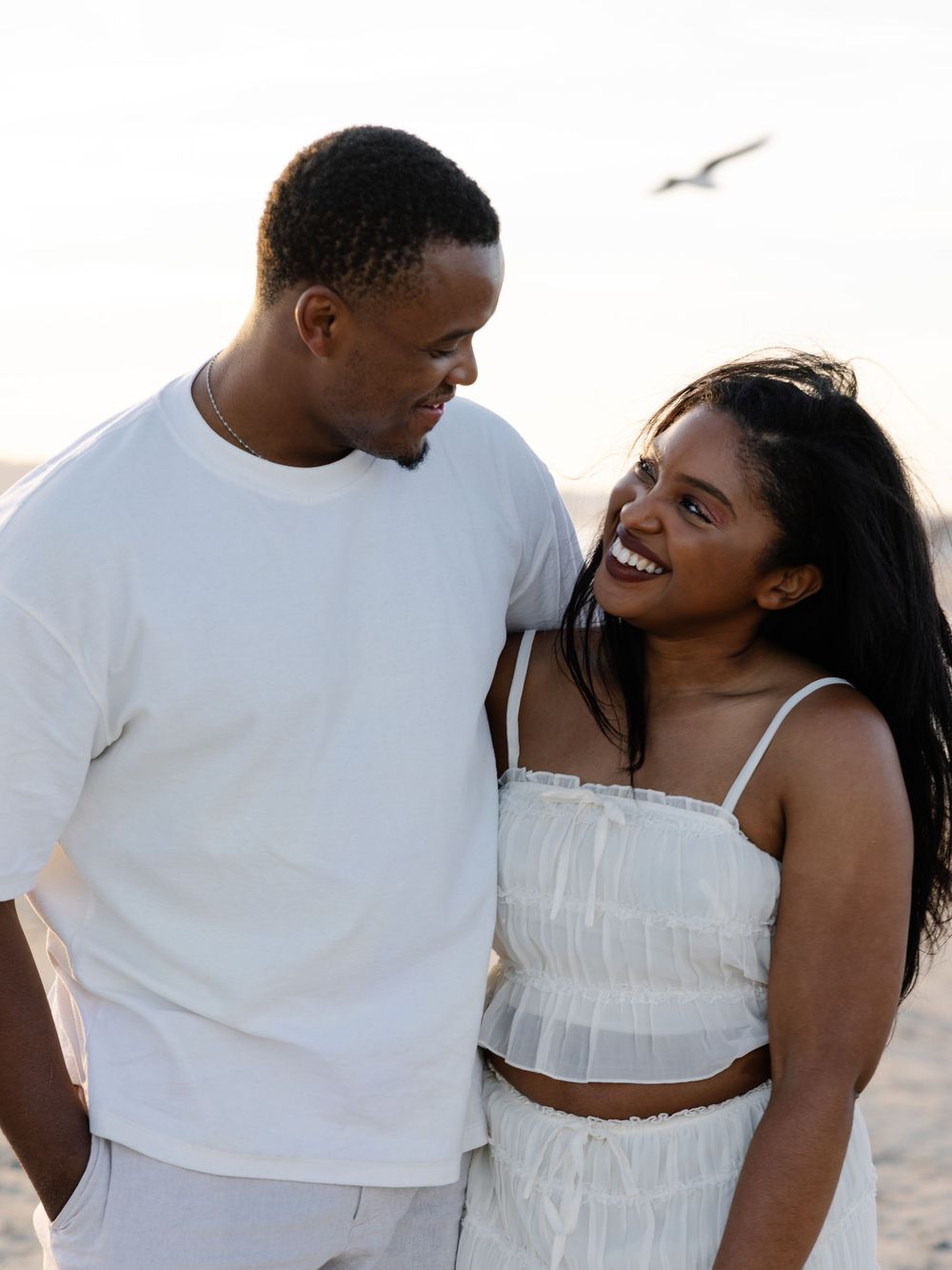 A happy Black couple in matching white outfits smiling at each other on a beach at sunset.