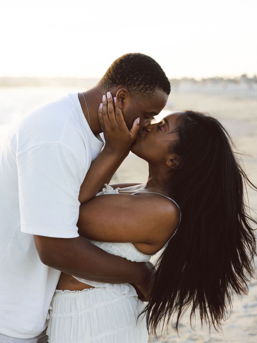 A couple sharing a romantic kiss on the beach at sunset, both dressed in white outfits.