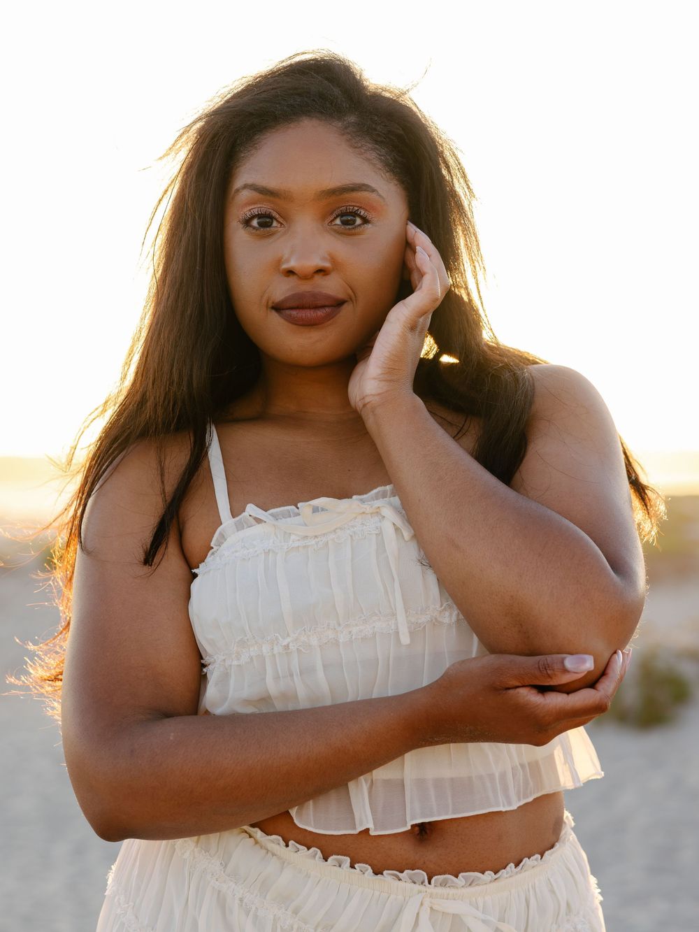 Young Black woman in white ruffled crop top poses gracefully on a beach at golden hour.