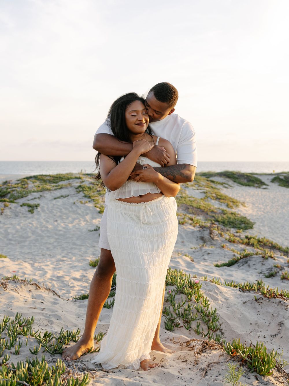 Couple embracing on sandy beach dunes at sunset, woman in white skirt and crop top, man in white t-shirt.