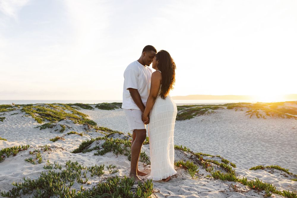 Couple embracing at sunset on white sand dunes, woman in white dress, golden hour beach photography.