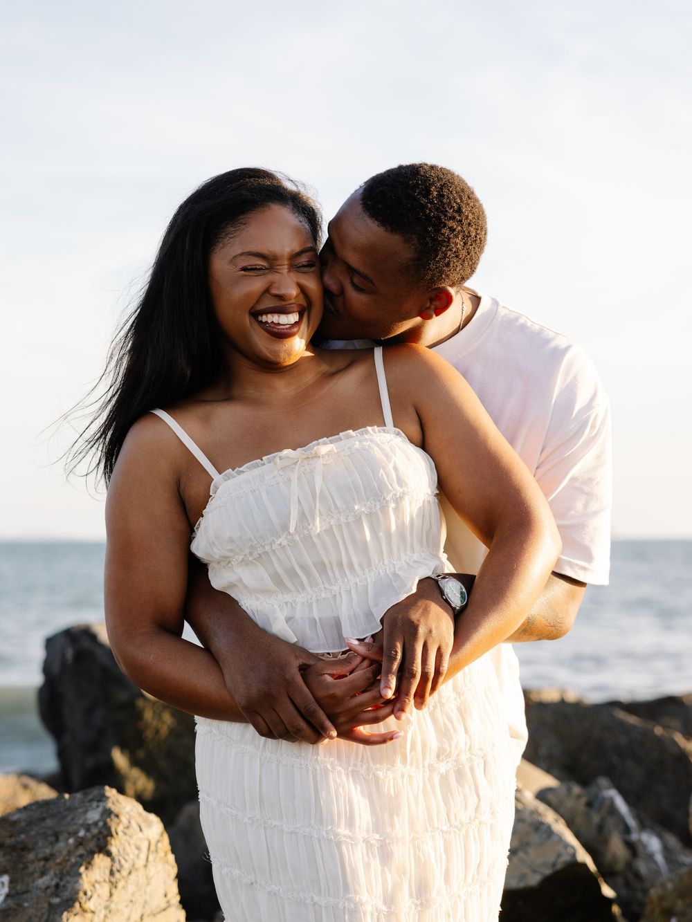 A joyful couple embraces on rocky shoreline, man kissing woman's cheek as she laughs in white dress.