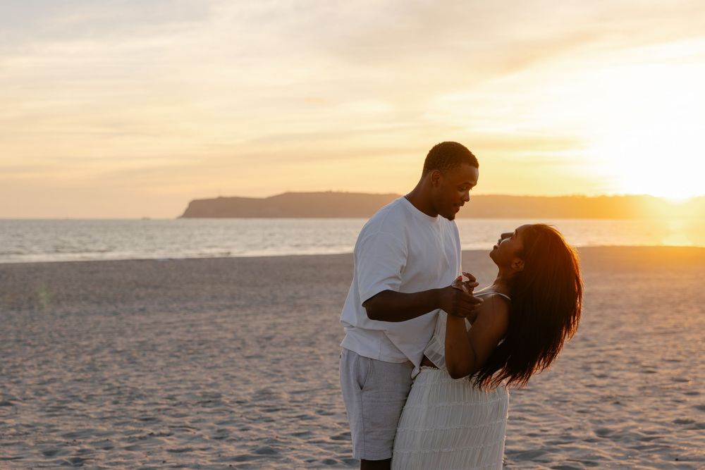 Couple dancing romantically on a sandy beach at golden sunset with ocean and hills in background.