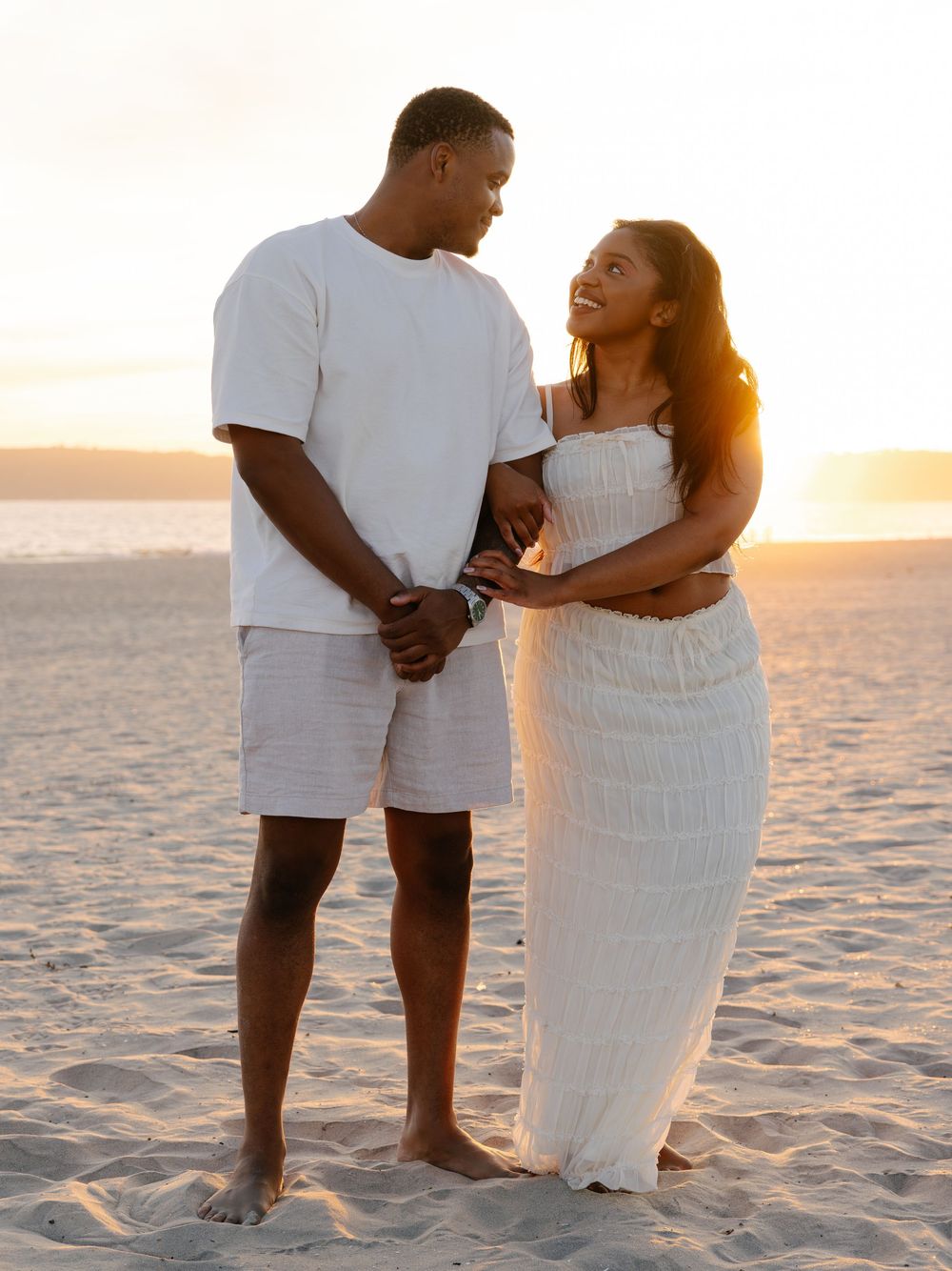Couple in white outfits holding hands on sandy beach at golden sunset, smiling at each other.