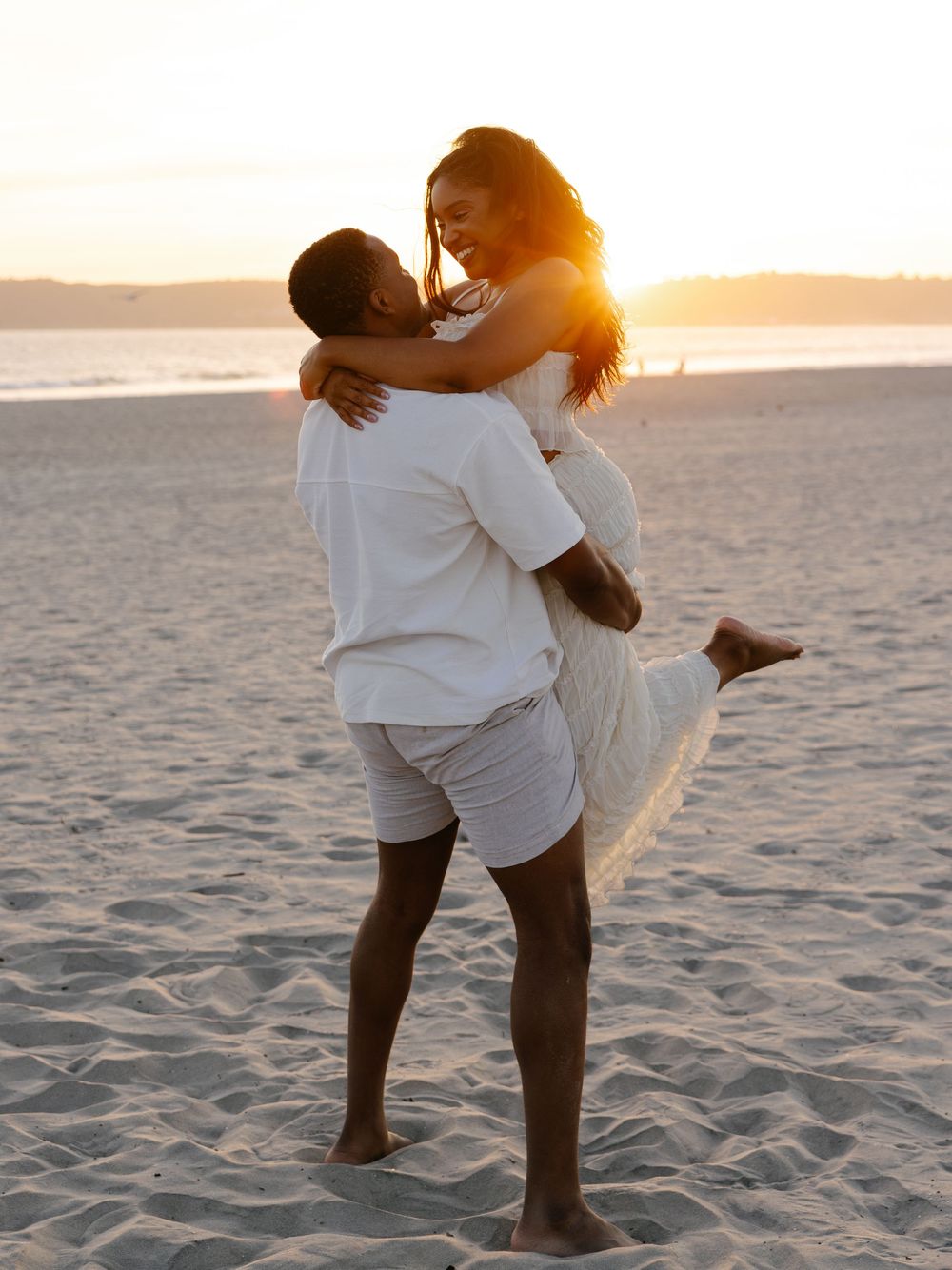 Couple in white outfits embracing on sandy beach at golden sunset, man lifting woman as they share a joyful romantic moment.