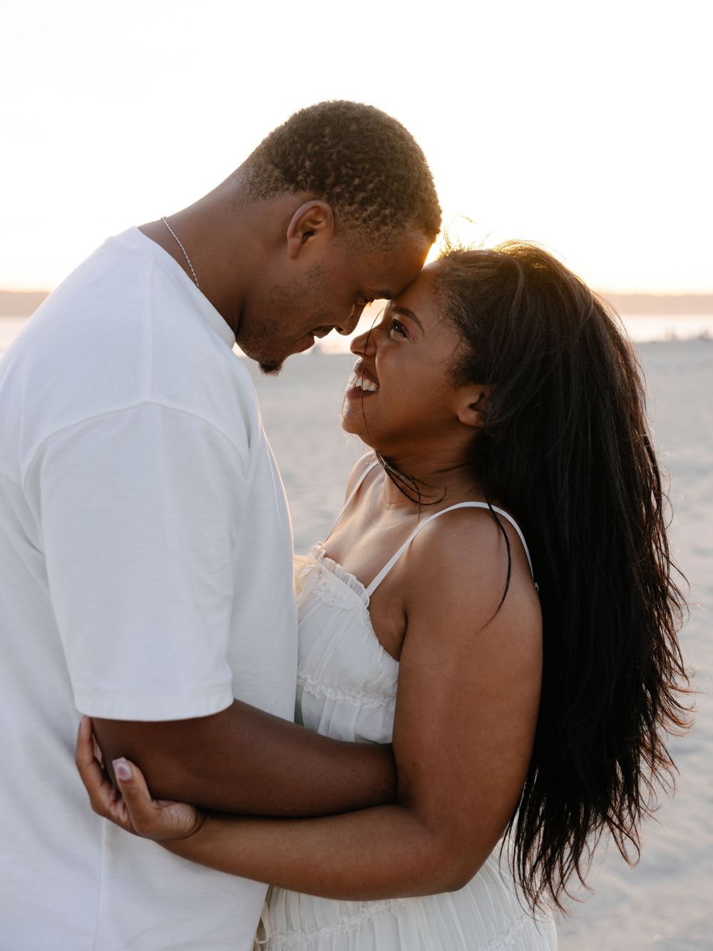 A joyful Black couple embraces on a beach at golden hour, touching foreheads and smiling in white outfits.