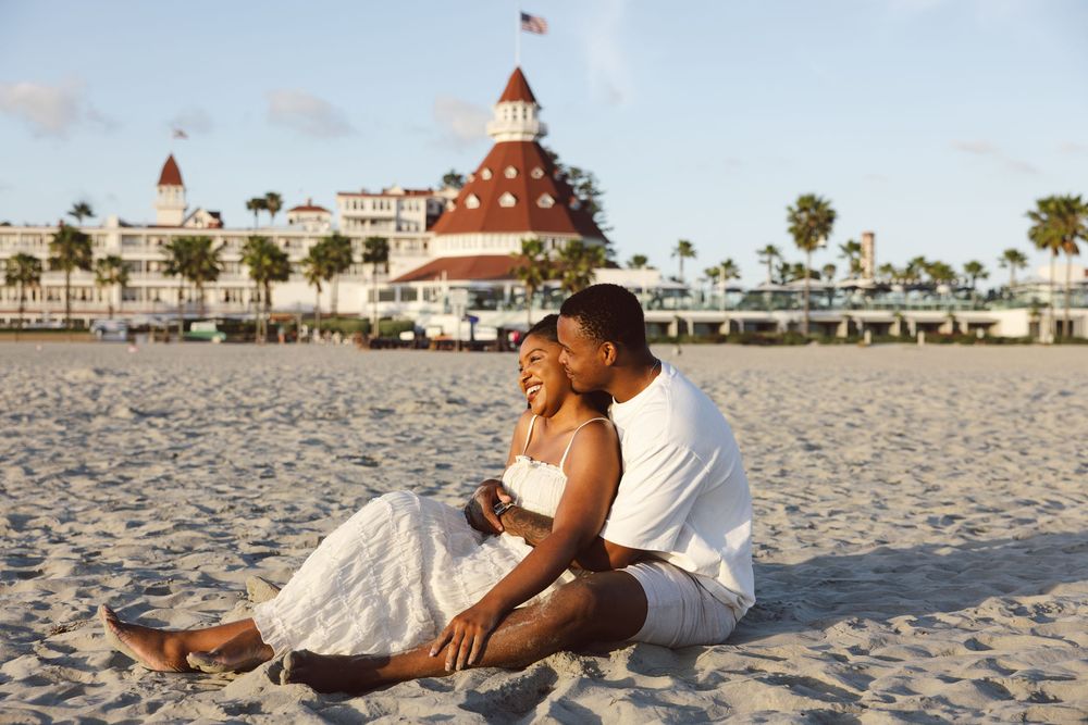 Couple sitting on sandy beach smiling, with iconic Hotel del Coronado red roof and palm trees in background.
