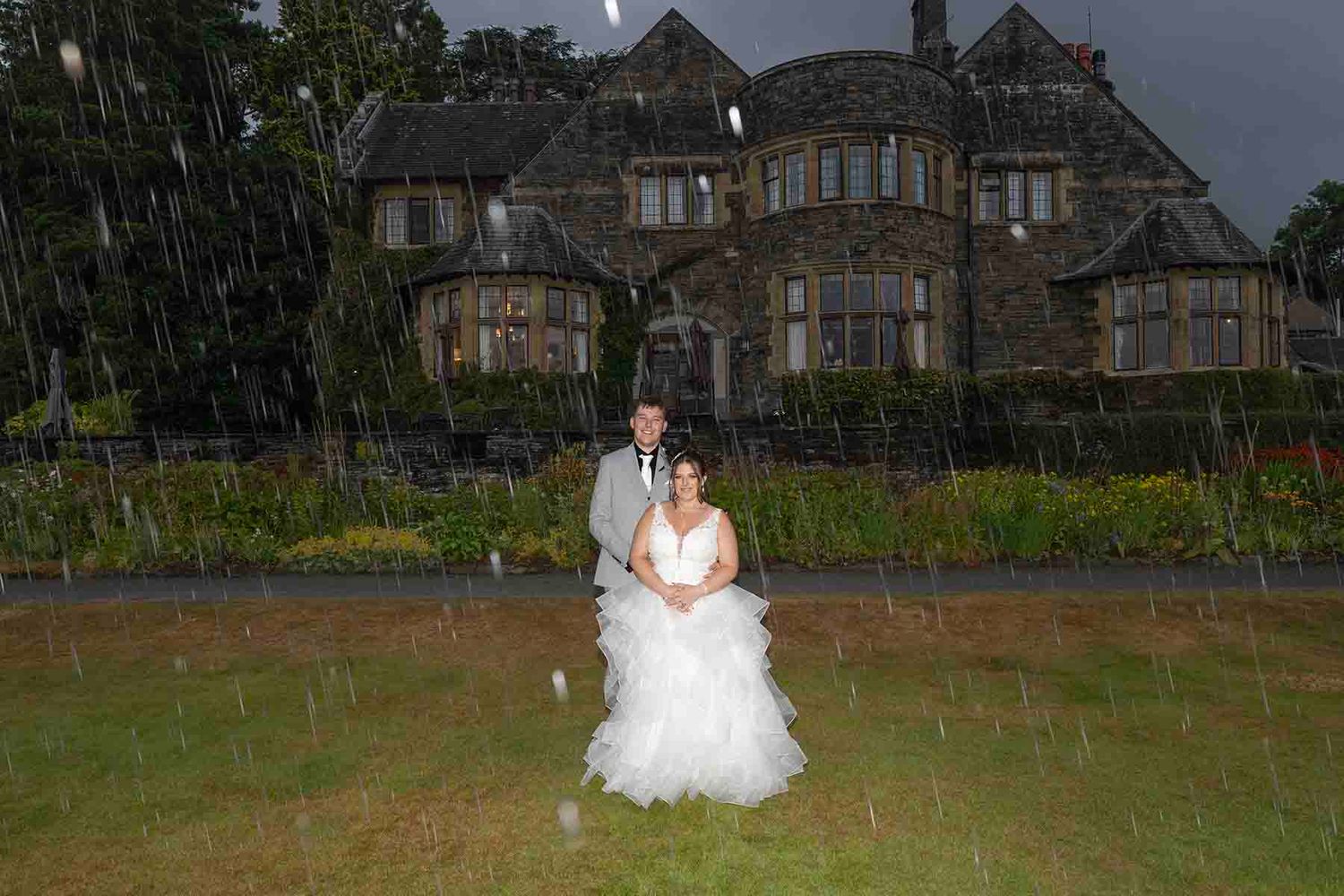 Bride and groom posing in the rain outside a grand stone manor house on their wedding day.