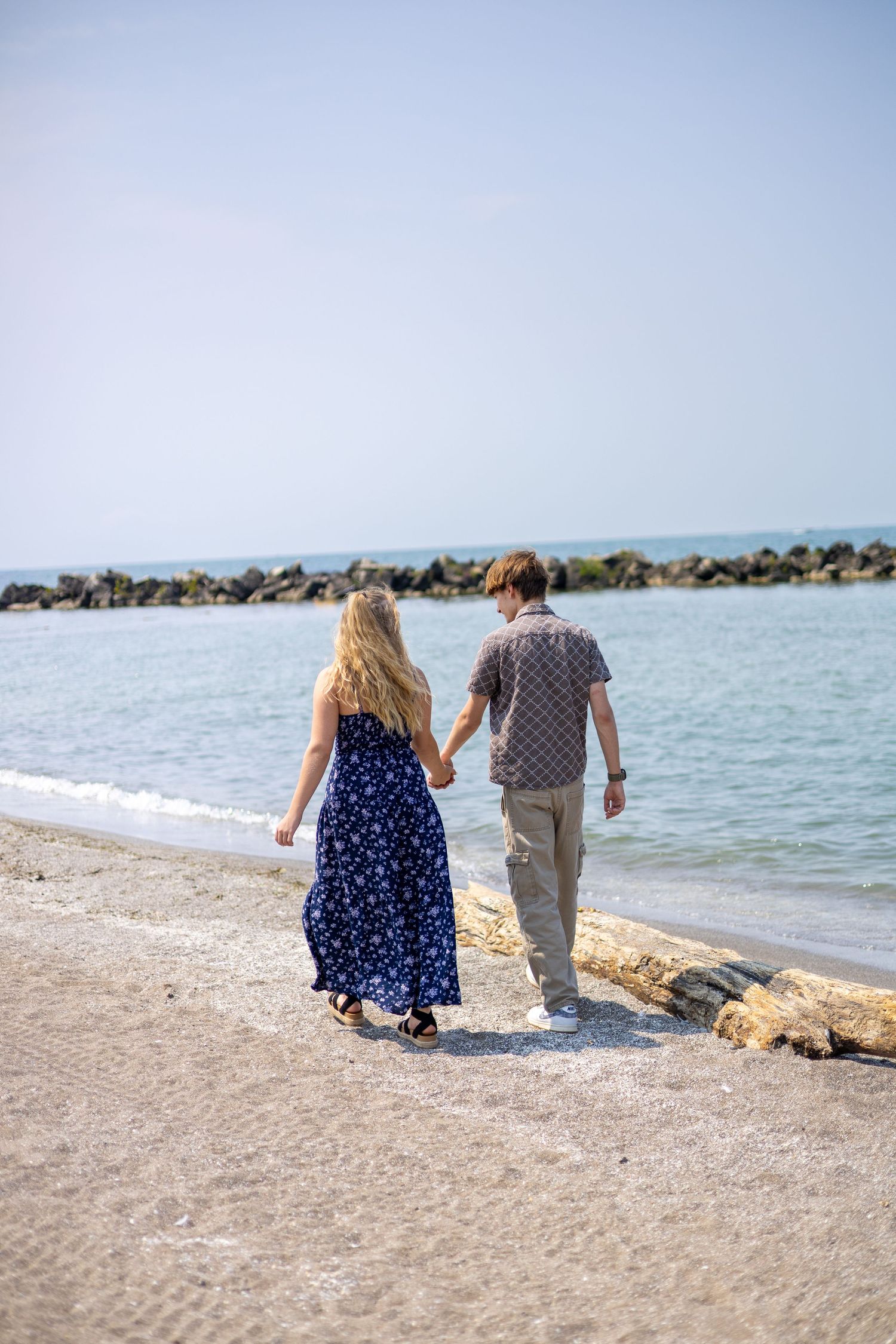 Couple walking hand in hand along a sandy beach toward a rocky breakwater on a sunny day.