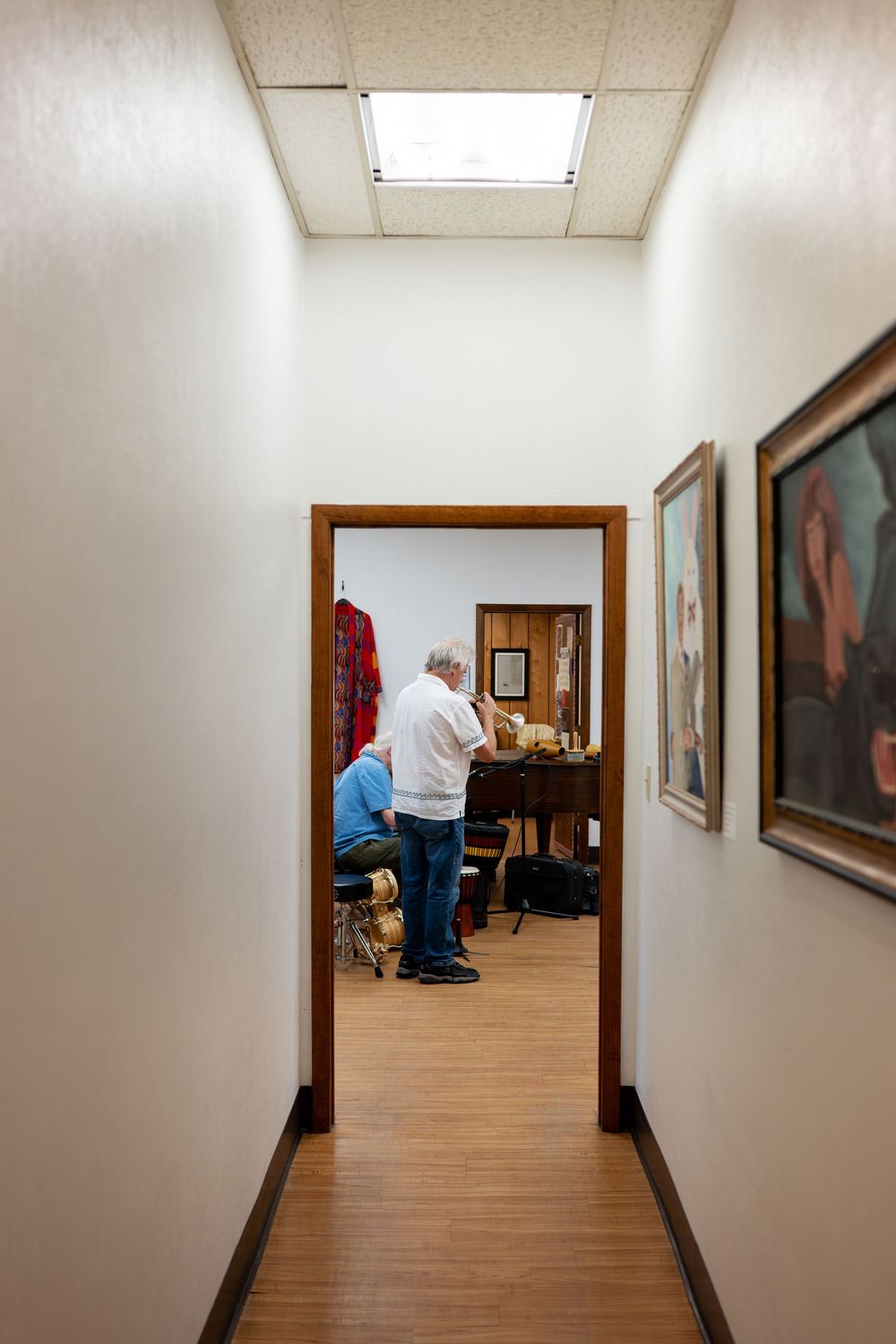 Narrow hallway with artwork on walls leading to a room where two people work at a desk in an art gallery.