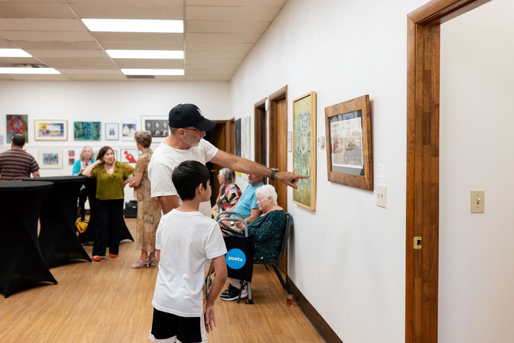 Adults and children viewing framed artwork on walls at an indoor gallery event with wood floors.