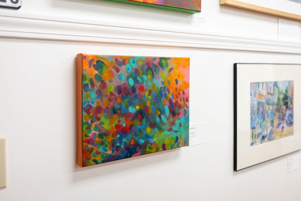 Colorful abstract painting with vibrant dots displayed on a white gallery wall next to framed artwork.