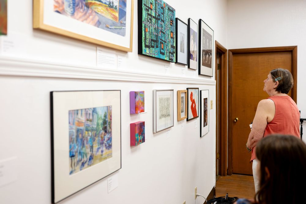 Woman viewing colorful framed artwork displayed on white gallery walls during an art exhibition.