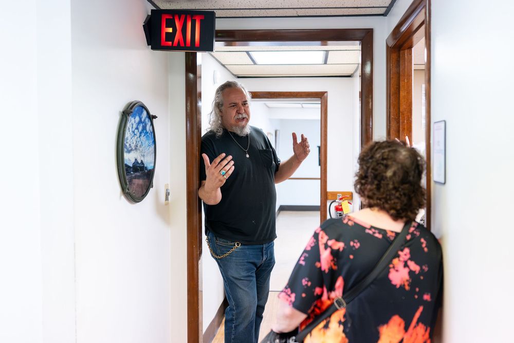 Man gesturing while speaking to a woman in a narrow hallway with small framed artwork.