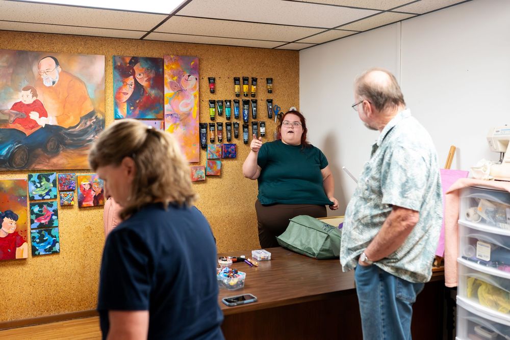 Three people discussing artwork displayed on a cork board wall in an office or art studio setting.