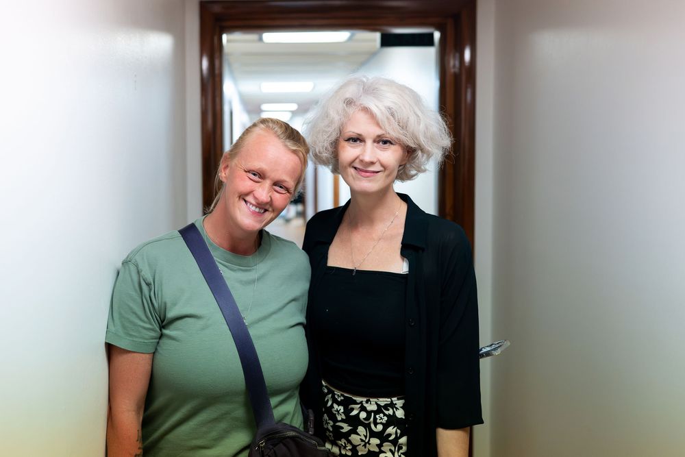 Two women smiling together in a hallway, one wearing a green t-shirt and the other in a black outfit.