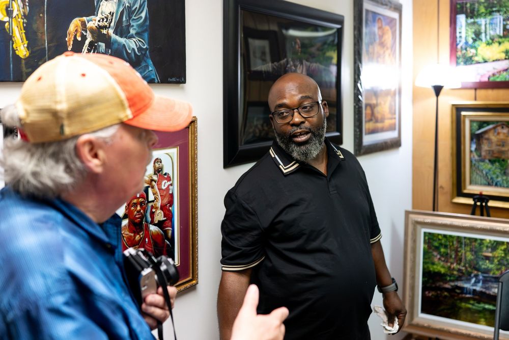Two men conversing in an art gallery surrounded by colorful framed paintings on the walls.