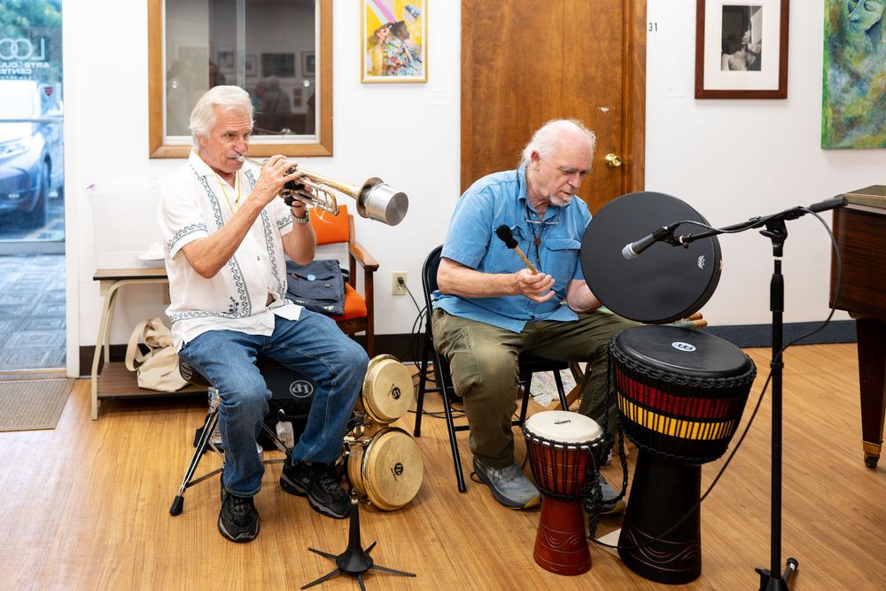 Two older men playing percussion instruments and trumpet in an art gallery setting.