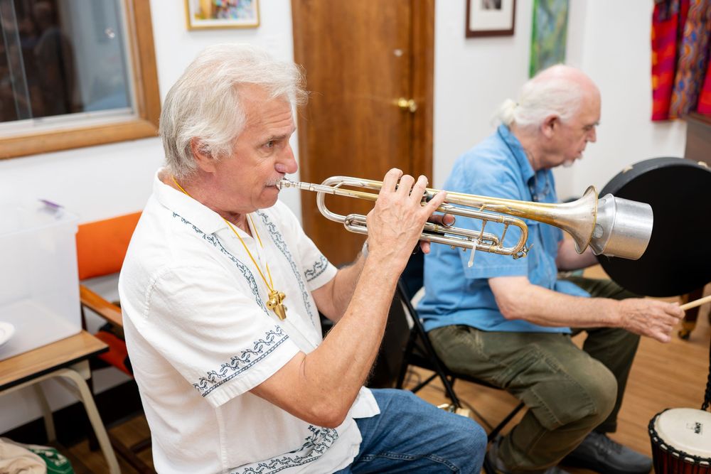 Two elderly men playing brass instruments, trumpet and tuba, seated indoors during a music rehearsal session.
