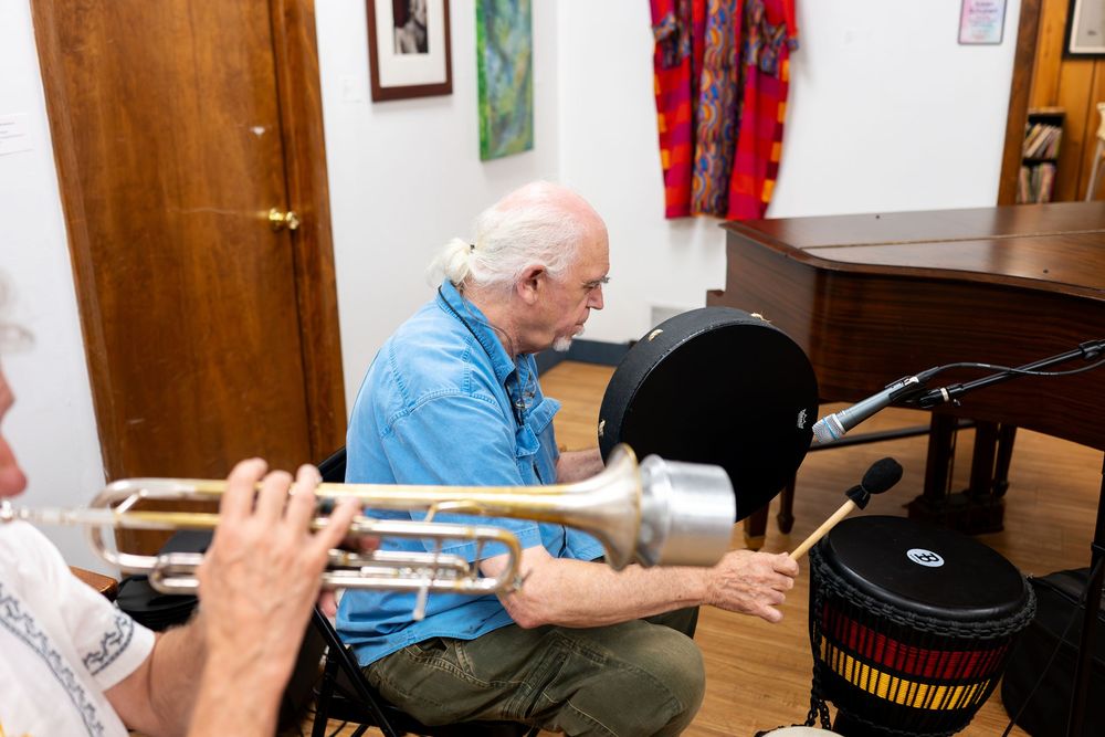Elderly man playing drums with a trumpet player in a cozy music room with colorful decor.