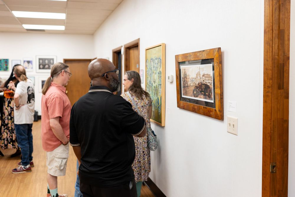 People viewing framed artwork on a wall at an indoor art gallery exhibition.