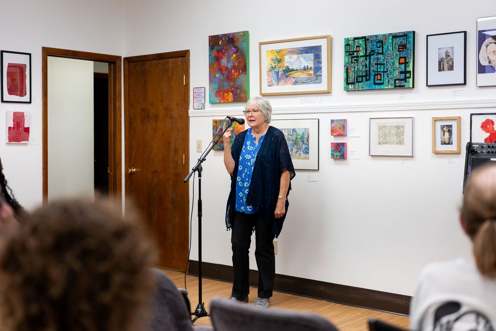 Woman speaking at microphone in art gallery with colorful framed artwork displayed on white walls.