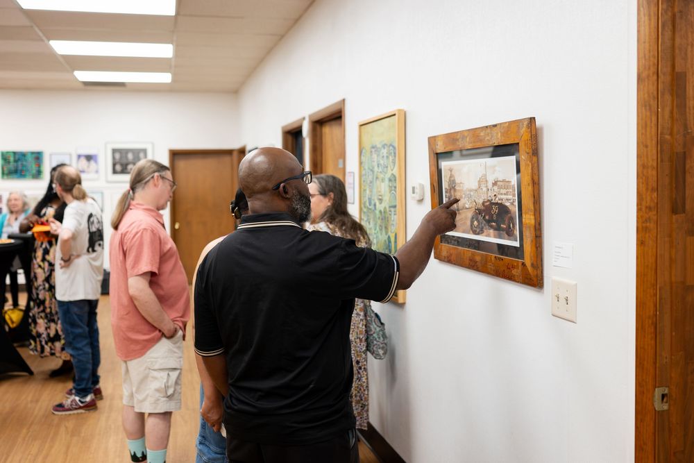 People viewing framed artwork on white gallery walls during an indoor art exhibition event.