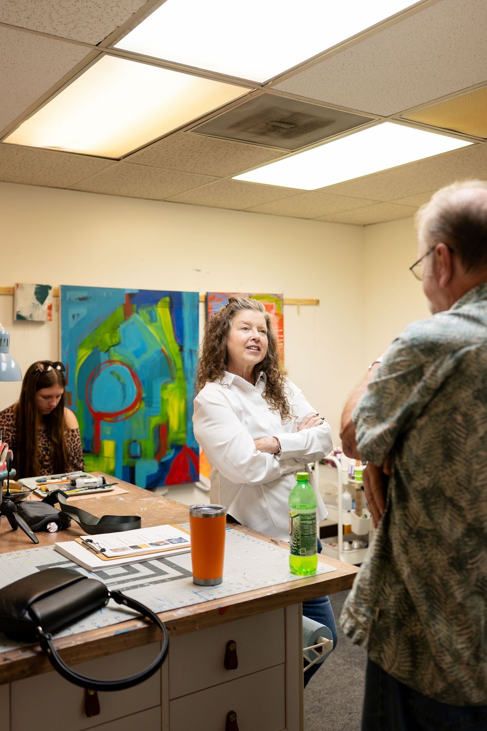 Woman with crossed arms discusses art in a studio with colorful abstract paintings displayed on the wall behind her.
