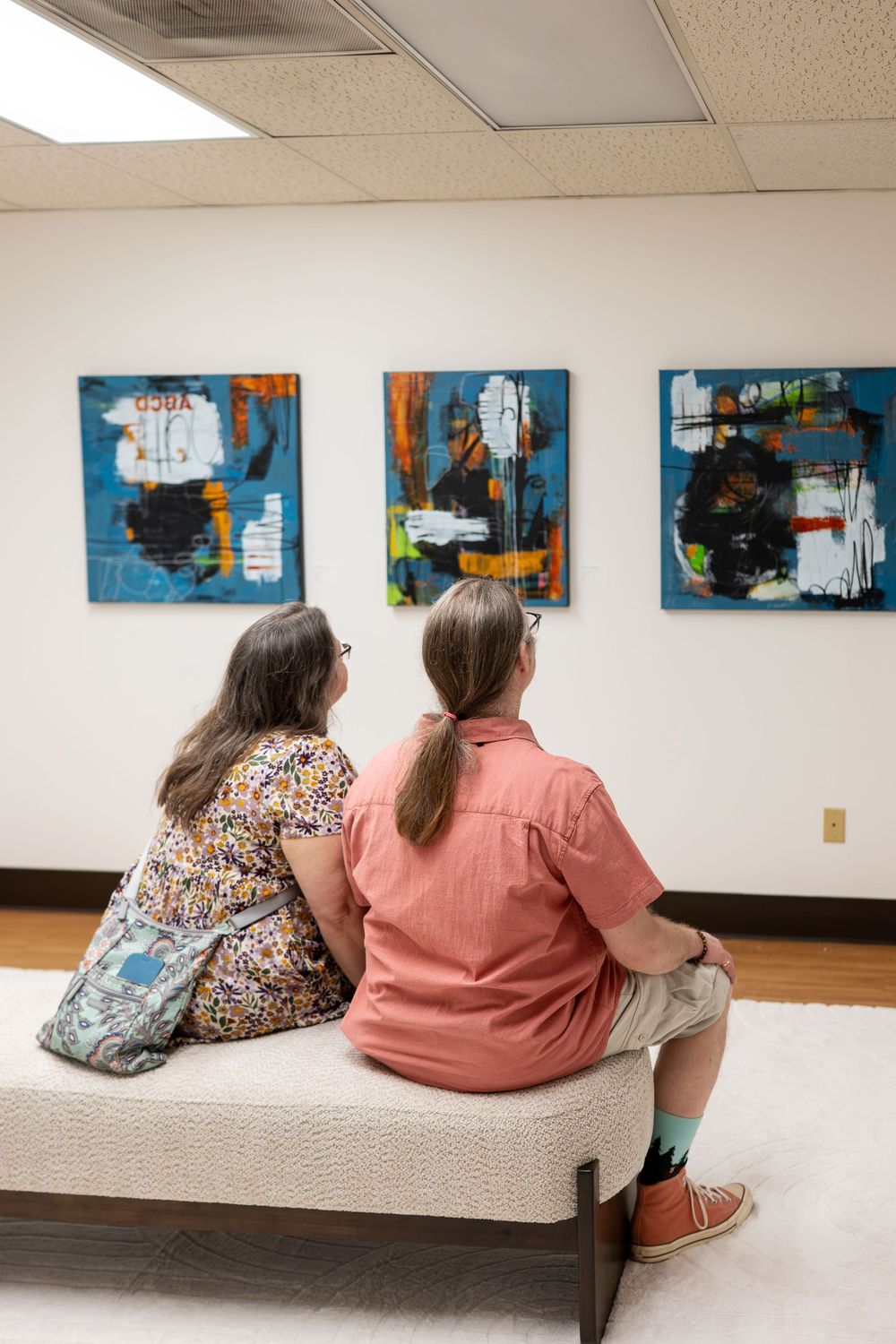 Two visitors sit on a bench viewing abstract blue and orange paintings in a modern art gallery.
