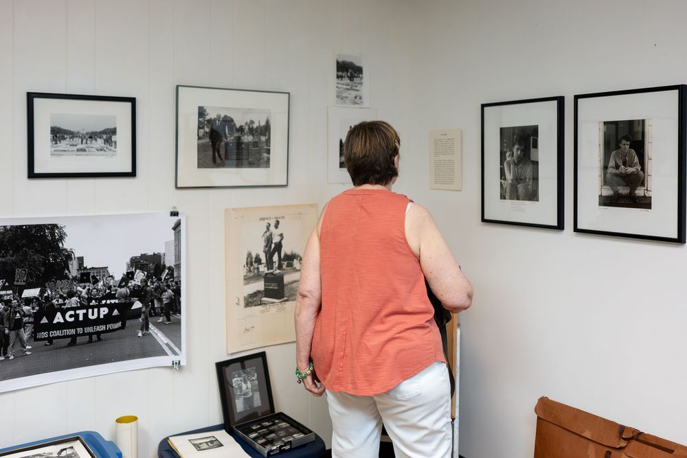 Person viewing framed black-and-white photographs and ACT UP memorabilia displayed on a white gallery wall.