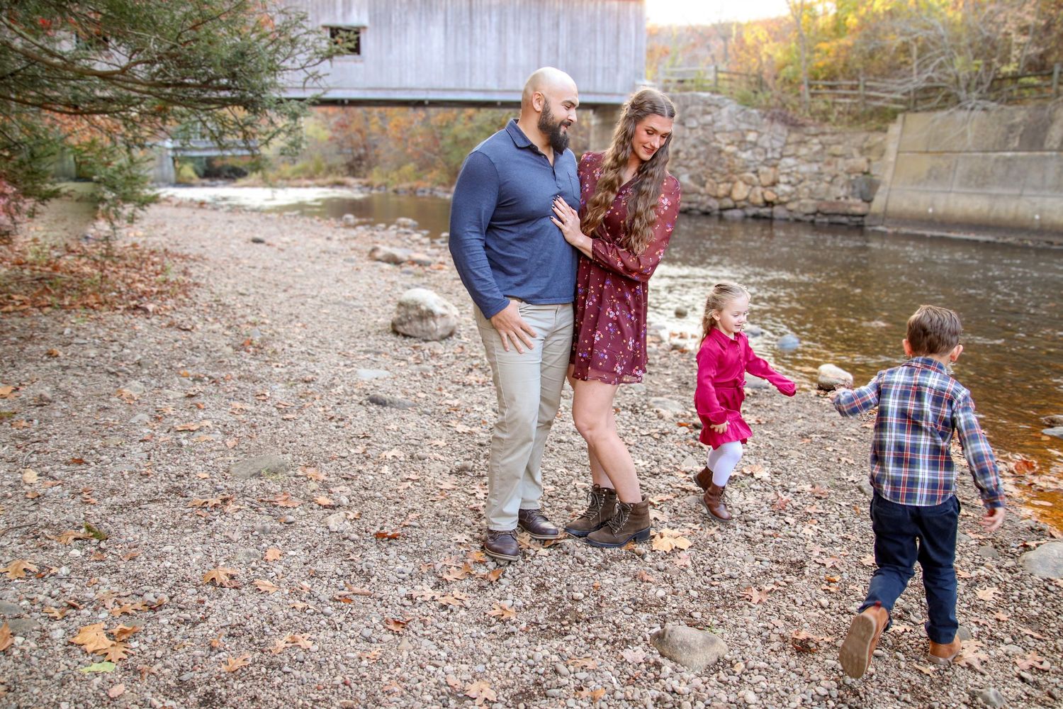 Family enjoying an autumn day by a rocky riverbank near a covered bridge with fall foliage during photo session in East Hampton, Connecticut