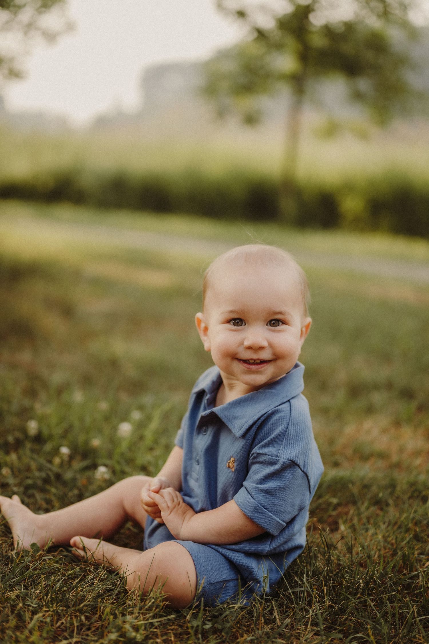 Smiling baby in blue romper sitting on green grass outdoors during golden hour.