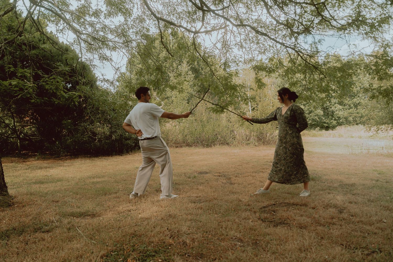 A couple playfully holds a stick together in a lush green park, dressed in casual summer attire.