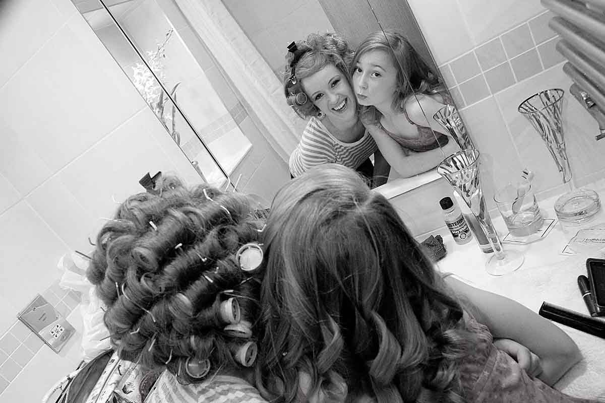 Black and white photo of two women smiling in a bathroom mirror, one with curlers in her hair.