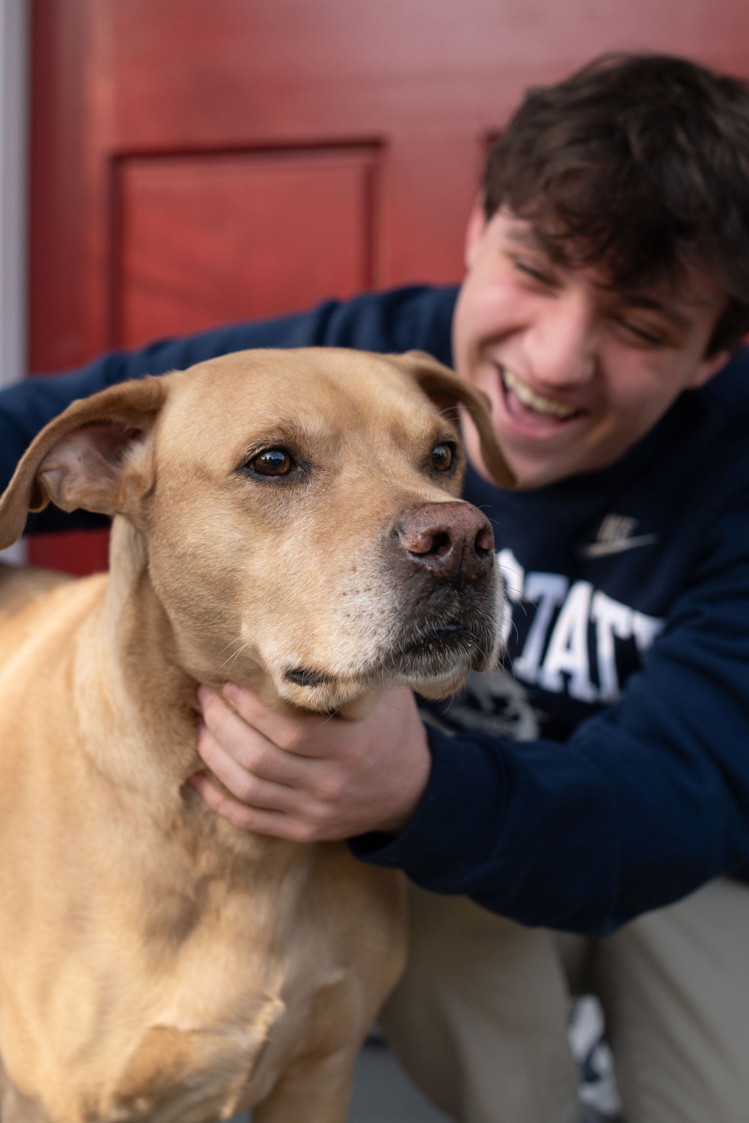 Smiling young man in a Penn State hoodie hugging a large tan dog near a red door.