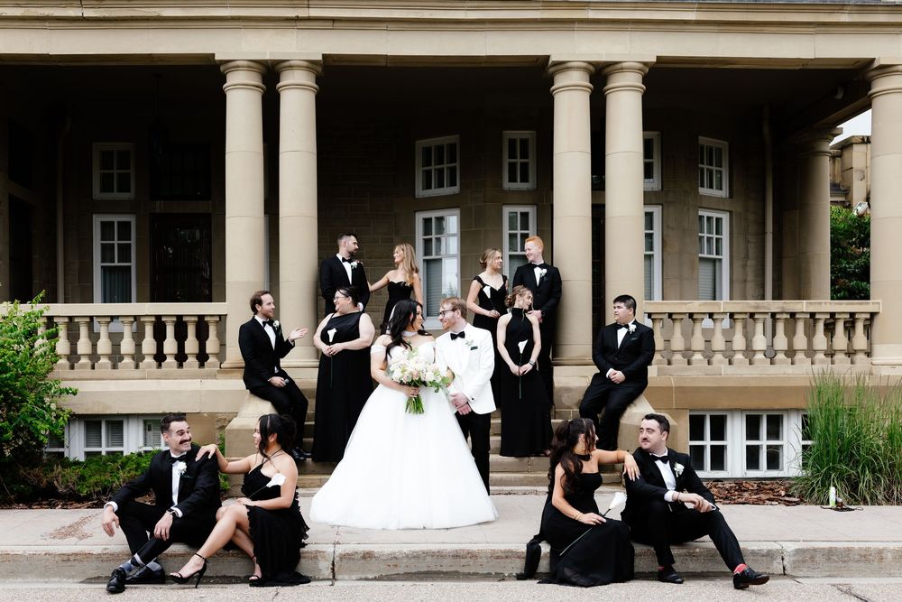 Wedding party in black and white formal attire posed on steps of elegant columned mansion with bride and groom centered.