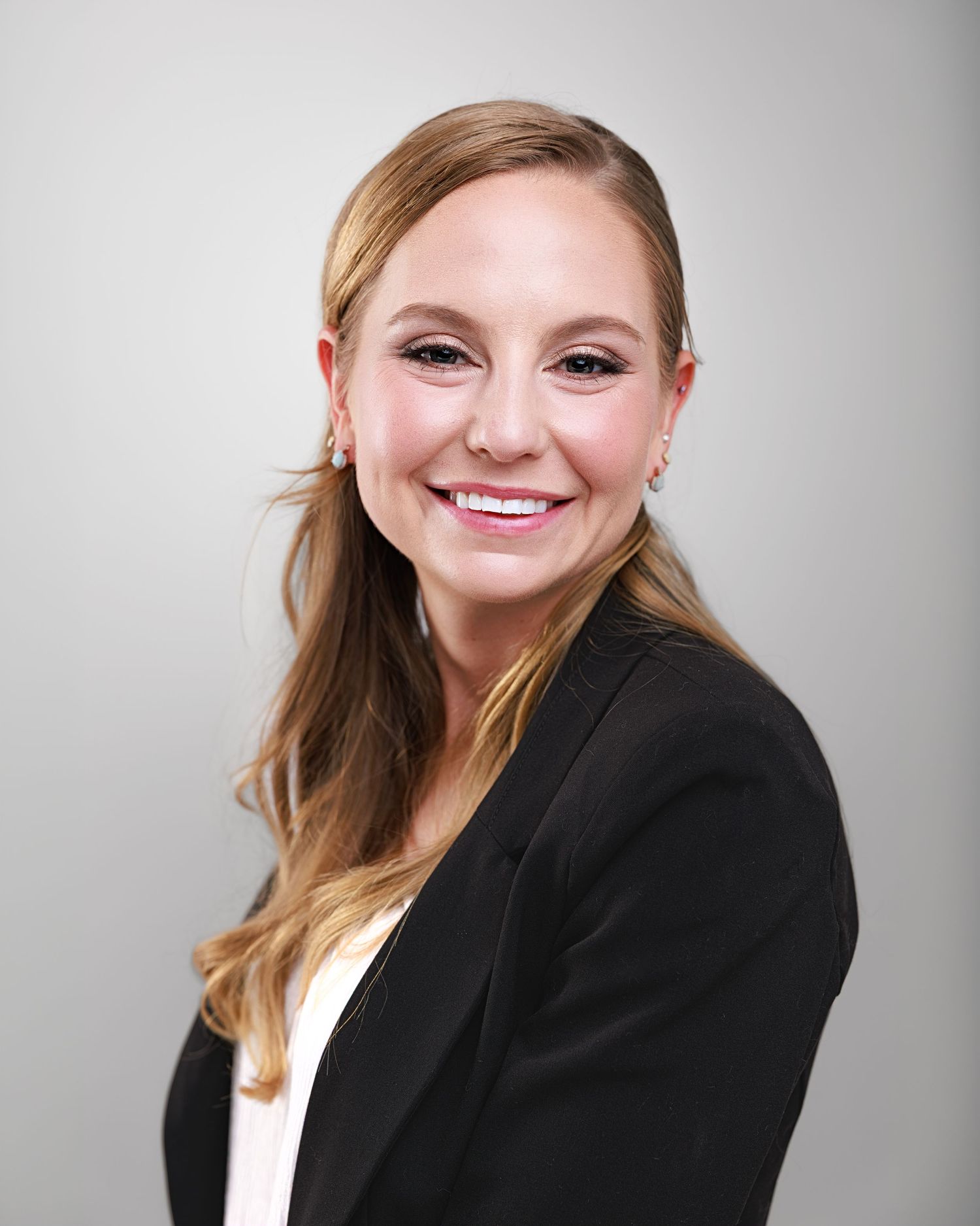 Professional headshot of a smiling blonde woman in a black blazer against a neutral gray background.
