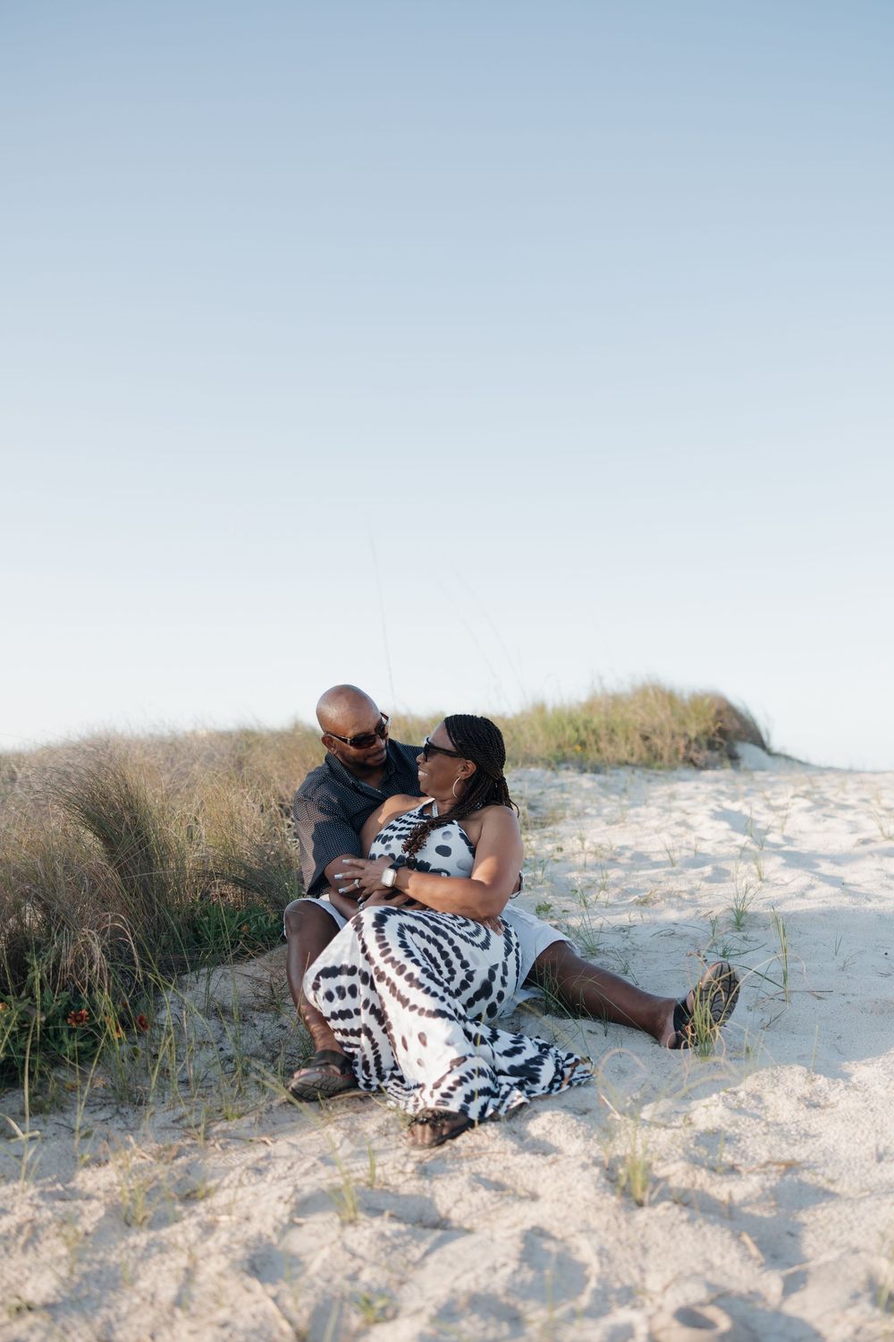 Couple sitting together on sandy beach dunes in matching black and white outfits at sunset.