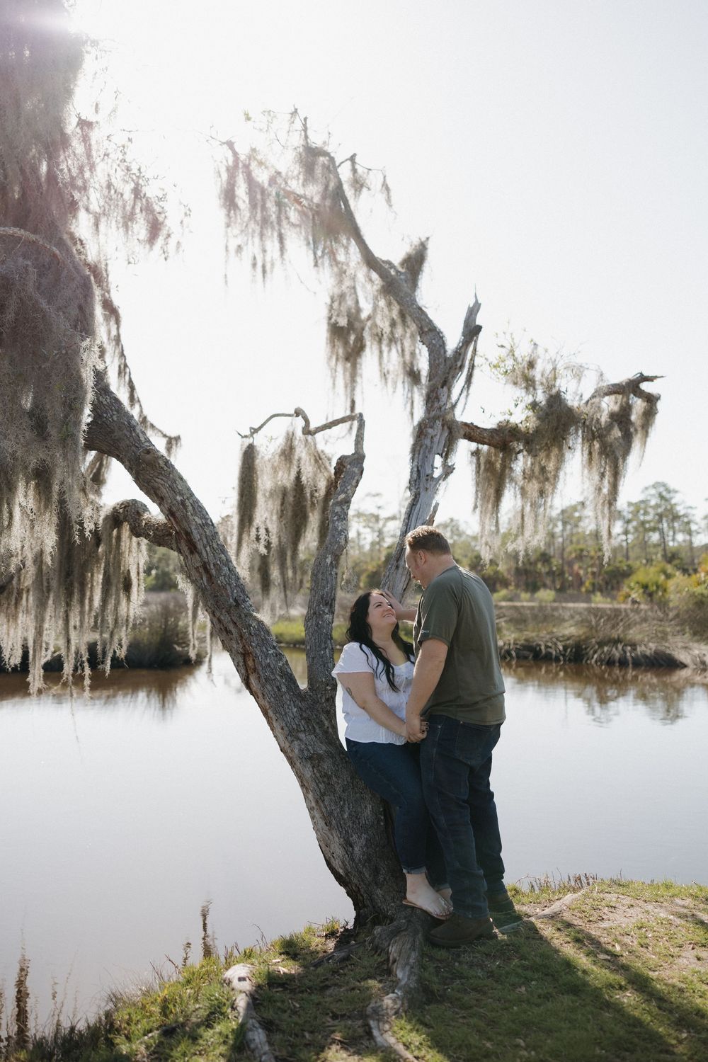Couple sharing a romantic moment under a moss-draped tree beside a tranquil lake.