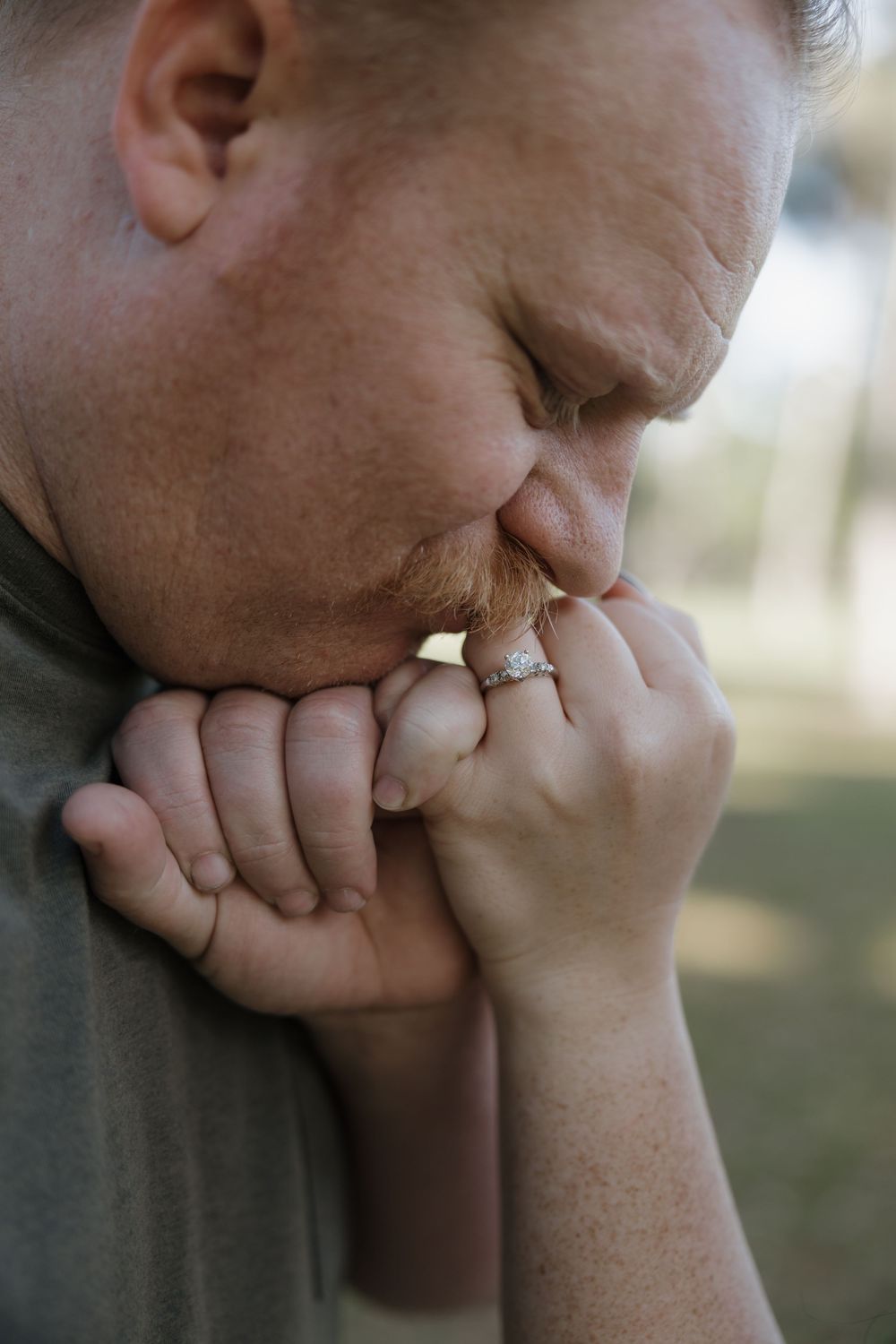 A man kisses a woman's hand tenderly, both wearing wedding rings, in an outdoor setting.