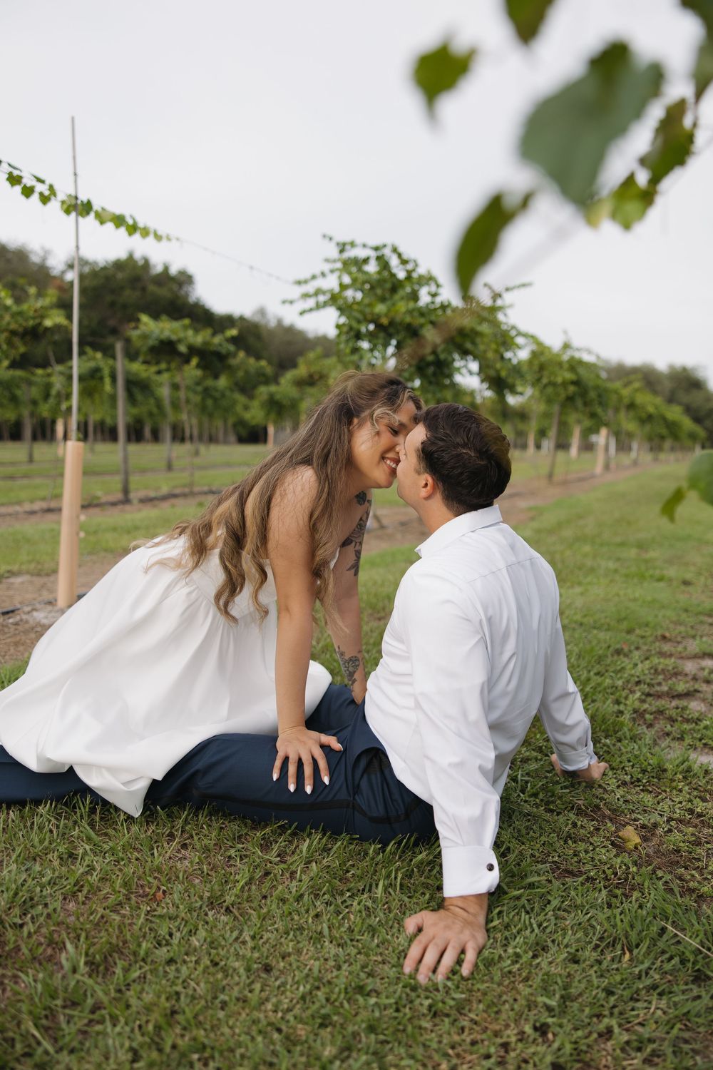 Couple sharing a kiss while sitting on grass in a vineyard, woman in white dress leaning toward man in white shirt.