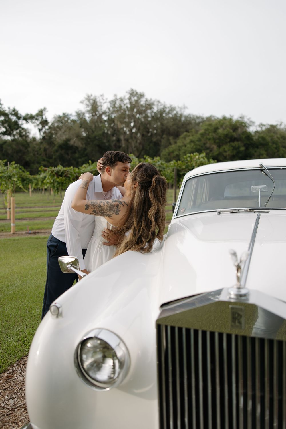 A couple shares a romantic kiss leaning against a classic white Rolls-Royce in a vineyard.