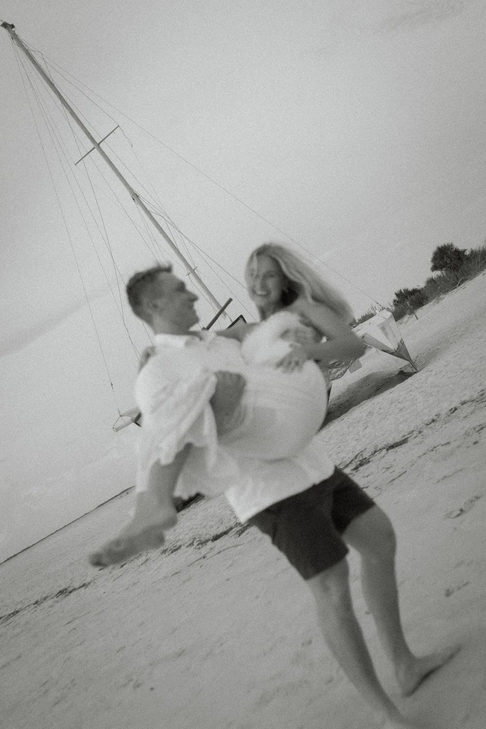 Black and white photo of a groom carrying his bride on a sandy beach with a sailboat in the background.