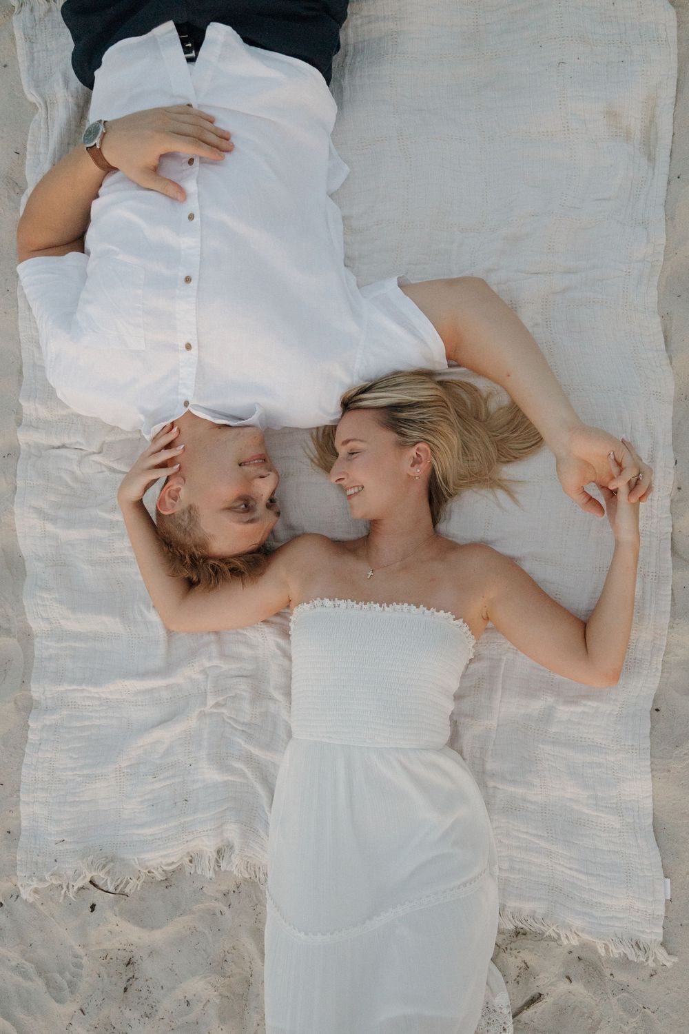 Couple in white outfits lying on a blanket on the beach, smiling at each other in a romantic moment.